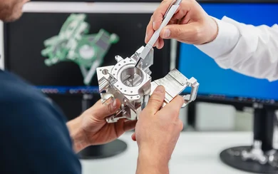 Zwei Hände halten ein Bauteil während eine dritte mit einem Kugelschreiber darauf zeigt