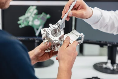 Zwei Hände halten ein Bauteil während eine dritte mit einem Kugelschreiber darauf zeigt